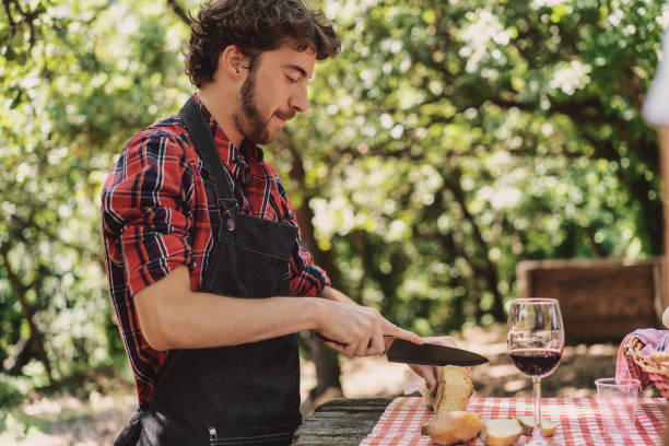 Bearded hipster man cutting the bread over a table in the countryside - young man preparing food for the picnic in the nature Bearded hipster man cutting the bread over a table in the countryside - young man preparing food for the picnic in the nature man beard plaid shirt stock pictures, royalty-free photos & images