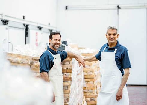 Portrait of two smiling butchers working in a meat packaging warehouse looking at camera. Two happy mature man wearing apron standing by a stack of packaged meet in a warehouse.