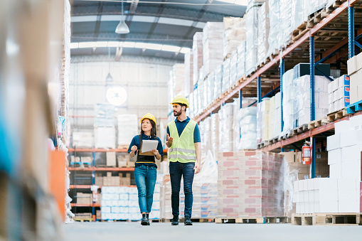 Male and female workers discussing over stock checklist in warehouse. Multi-ethnic colleagues are working together at distribution warehouse, walking by the racks and looking the stock levels.