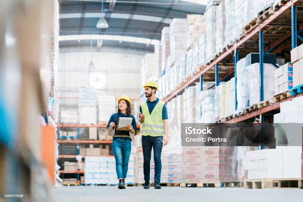 Two employees checking inventory on warehouse racks Male and female workers discussing over stock checklist in warehouse. Multi-ethnic colleagues are working together at distribution warehouse, walking by the racks and looking the stock levels. Freight Transportation Stock Photo