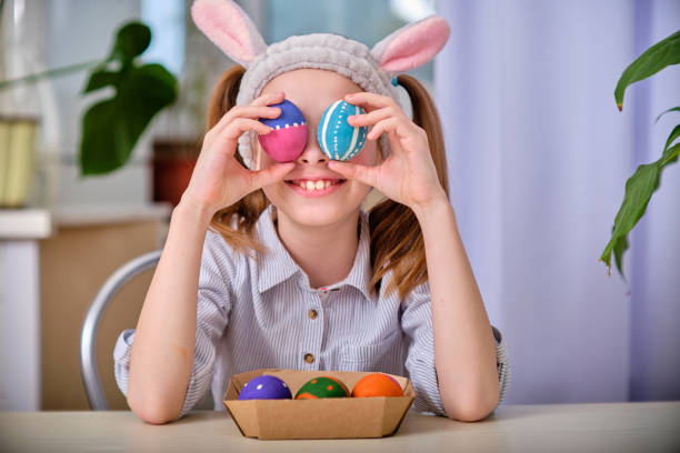 ein positives verspieltes mädchen in hasenohren hält in der heimischen küche bunte eier in den händen. frohe ostern. osterhintergrund - easter easter bunny fun humor stock-fotos und bilder