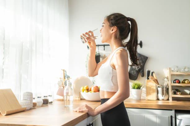 mujer hermosa asiática en ropa deportiva bebe agua después del ejercicio en casa. joven y sedienta de deporte activo toma un sorbo de mineral natural limpio en la taza después del entrenamiento para el cuidado de la salud en la cocina de la casa. - cuidado corporal y belleza fotografías e imágenes de stock