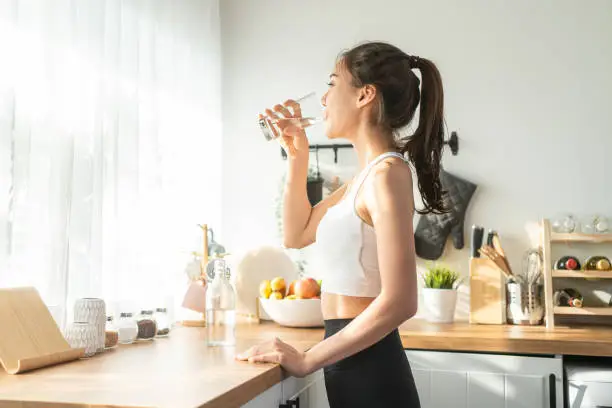 Photo of Asian beautiful woman in sportswear drink water after exercise at home. Young thirsty active sport girl takes a sips of clean mineral natural in cup after workout for health care in kitchen in house.