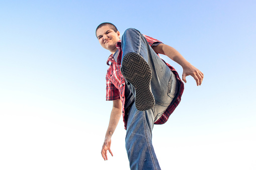 A young woman with a shaved head lifts her foot over the camera, as if she is about to stomp on it.  The background is a vibrant blue sky.