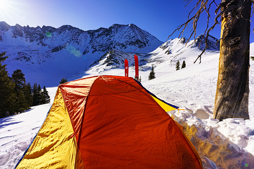 High Mountain Ski Camping - Ski mountaineering at high altitude with colorful orange tent perched on mountain ridge with skis and rugged mountains in background.