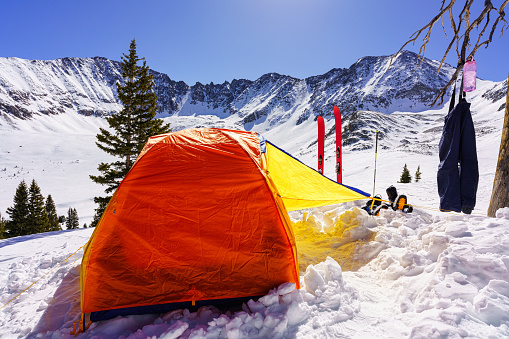 High Mountain Ski Camping - Ski mountaineering at high altitude with colorful orange tent perched on mountain ridge with skis and rugged mountains in background.