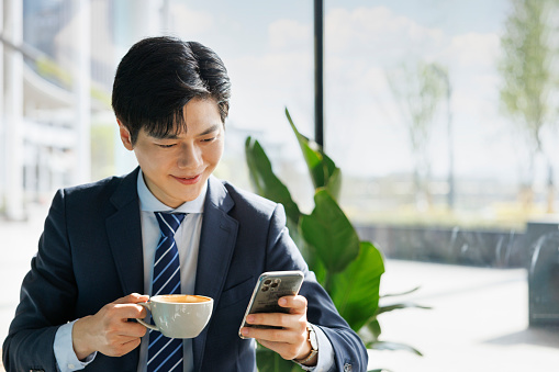 A young asian male businessman, about 30 years old, wearing a gray suit, sitting in a bright cafe, drinking coffee and using a mobile phone.
