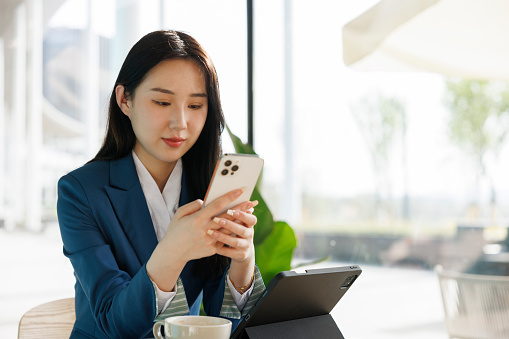 A young asian female businessman, about 25 years old, wearing a blue suit, sitting in a bright coffee shop and using a mobile phone to contact work.