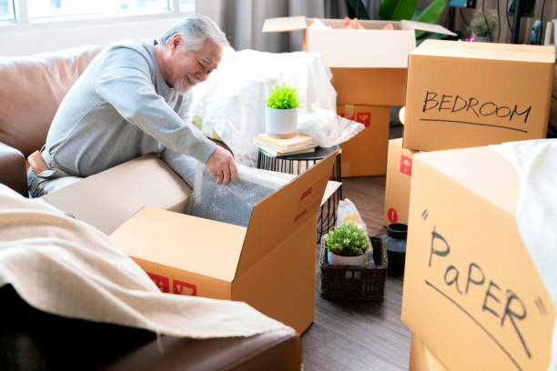 Horizontal medium shot of adult elder senior asian man male white beard sitting on floor preparing for moving to new house packing things into boxes and taping them in living room,home moving ideas concept foto mediana horizontal de anciano adulto mayor asiático hombre de barba blanca sentado en el suelo preparándose para mudarse a una nueva casa empacando cosas en cajas y pegándolas en la sala de estar, concepto de ideas de mudanza de casa
