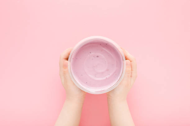 밝은 분홍색 테이블 배경에 베리 요구르트의 플라스틱 컵을 들고있는 아기 소녀 손. 파스텔 색상. 근접 촬영. 시점 촬영. 아이들은 건강하고 맛있는 음식. 하향식 보기. - yogurt container 뉴스 사진 이미지