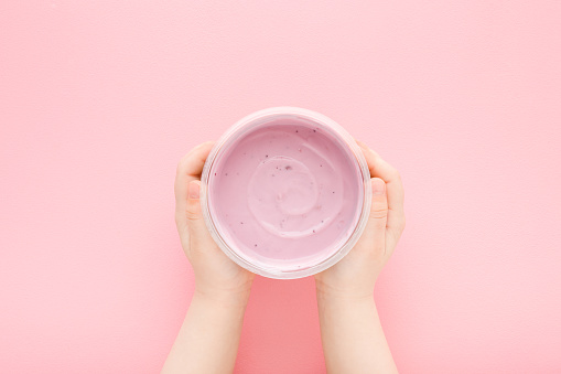 Baby girl hands holding plastic cup of berry yogurt on light pink table background. Pastel color. Closeup. Point of view shot. Children healthy, tasty food. Top down view.