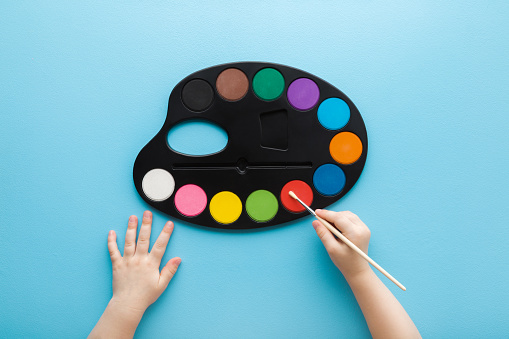 Baby hand holding paint brush and using colorful palette on light blue table background. Pastel color. Closeup. Point of view shot. Top down view.