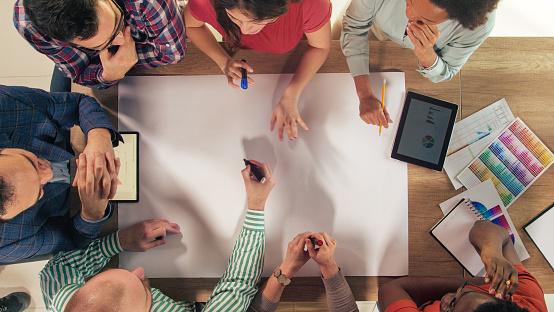 Directly above shot of diverse group of entrepreneurs sitting around a conference table, big blank paper in the middle, creating a new business plan.