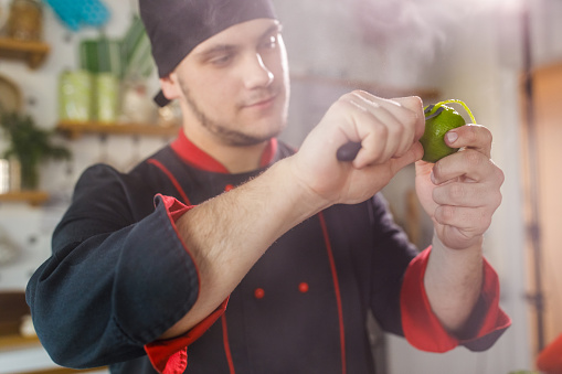 Selective focus shot of young smiling chef peeling a lime, to add the peel as garnish on top of a dish he prepared.