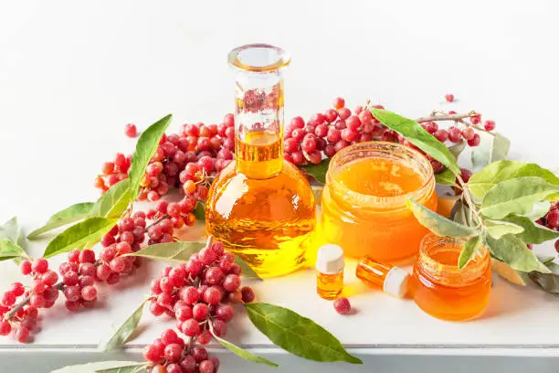 Natural products for healthy eating. Glass bottle with oil, glass jars with honey, branches of red sea buckthorn berries in a white wooden table against a light gray background, top view.
