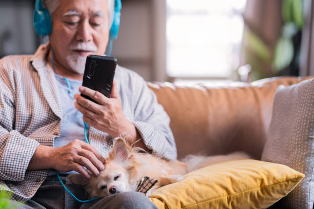 犬の親友のペットとヘッドフォンを着けたアジアの老人がソファに座り、自宅の居心地の良いリビングルームで休みながら一緒に歌う音楽を聴いている - man made structure high definition television domestic room living room ストックフォトと画像