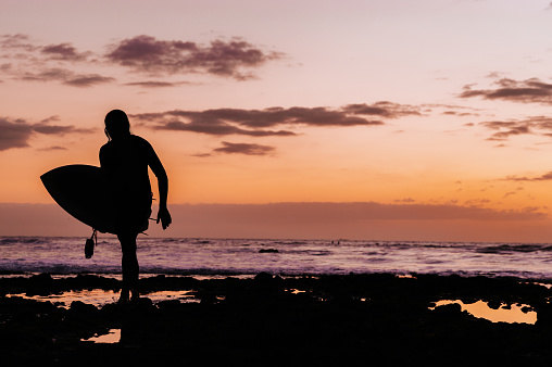 A silhouette of a man carrying a surfboard on his head and supporting it with his hands, he is getting ready to surf. He is at Kuta beach in Bali, Indonesia at dusk.