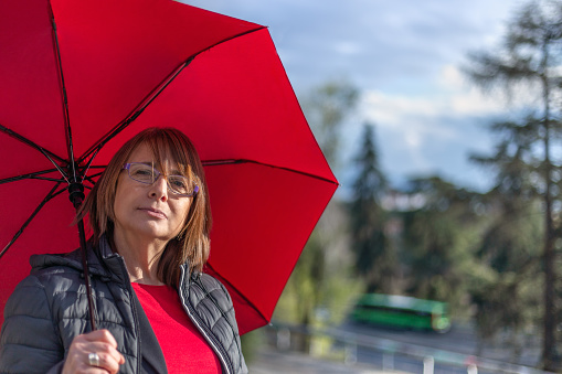 Madrid, Spain, March 7 2022. Middle-aged woman with glasses under red umbrella looking at camera in an instant of sunshine from a stormy day. In the background, unfocused, green bus moving at high speed. Concepts: Weather, meteorology