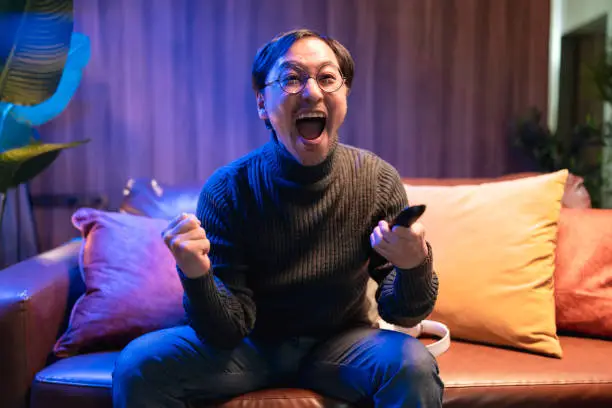 Photo of Asian adult man in front of the television watches a game of sports sitting on the couch .excited man watching football game hand rising up celebrating scoring goal screaming happy cheering at home