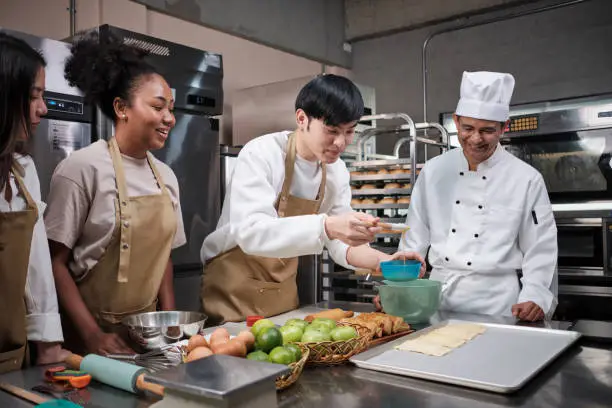 Cuisine course, senior male chef in uniform teaches young cooking class students, brushes pastry dough with eggs cream, prepares ingredients for bakery foods, fruit pies in stainless steel kitchen.