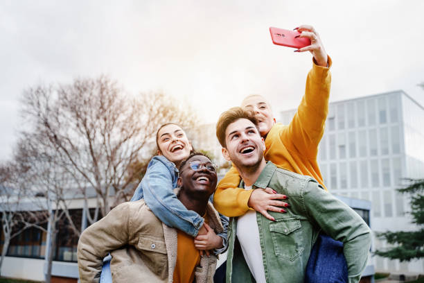 grupa happy young university student friends piggyback, bawiąca się śmiechem i robieniem selfie zdjęciem telefonem komórkowym. skoncentruj się na afroamerykaninie - mobile phone group of people photographing teenager zdjęcia i obrazy z banku zdjęć