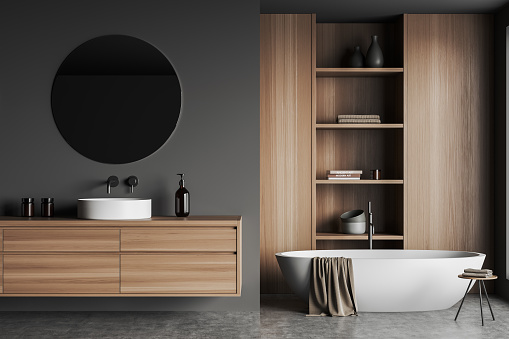 Dark bathroom interior with sink and mirror, tub on grey concrete floor, table with bathing accessories and shelf with decoration. Washing room in hotel with gels and towel, 3D rendering