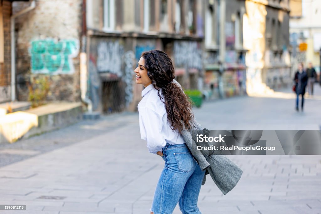 Young woman walking.Woman smiling while walking outdoors on the street. Happy young adult woman smiling with teeth smile outdoors and walking on city street.Beautiful brunette young woman wearing white shirt and a gray coat and walking on the street. 25-29 Years Stock Photo