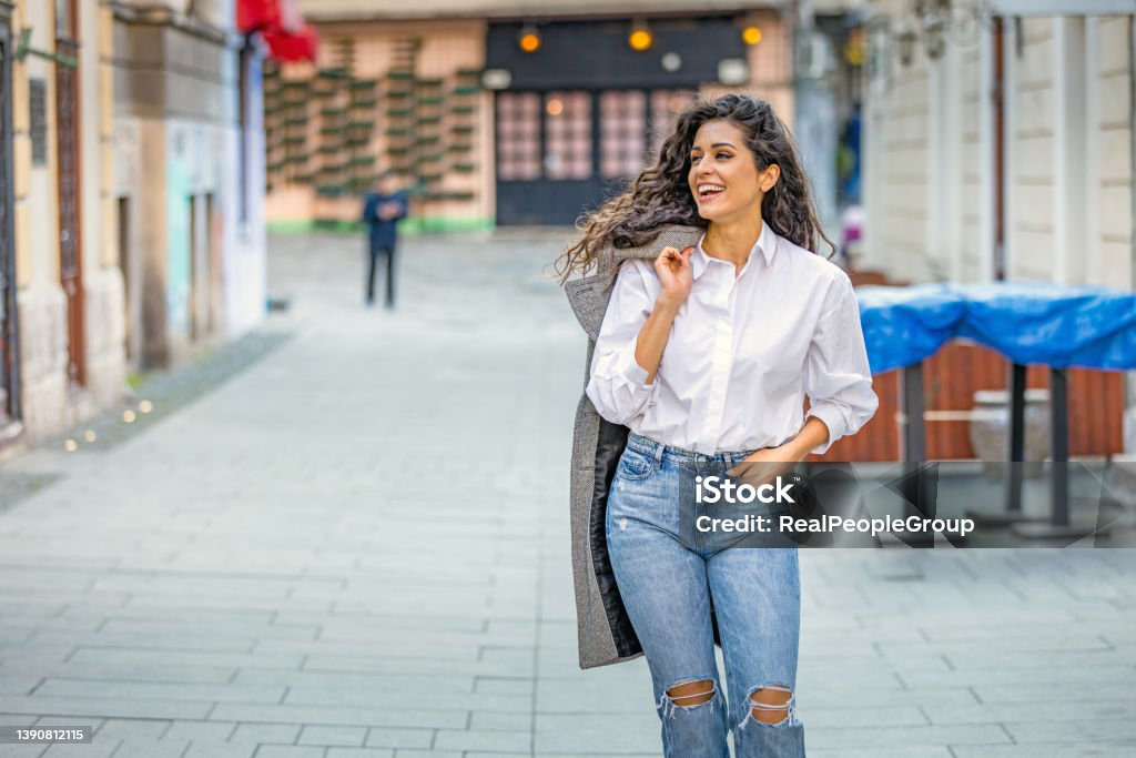 Young woman walking.Woman smiling while walking outdoors on the street. Happy young adult woman smiling with teeth smile outdoors and walking on city street.Beautiful brunette young woman wearing white shirt and a gray coat and walking on the street. Care Stock Photo