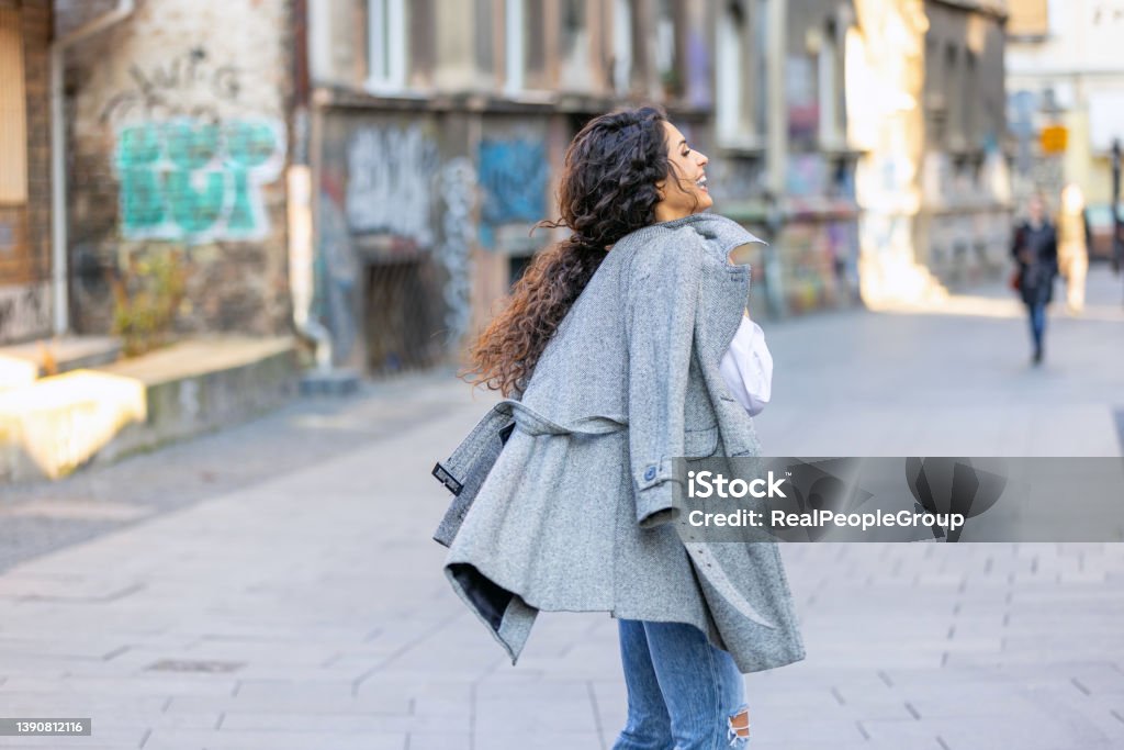 Young woman walking.Woman smiling while walking outdoors on the street. Happy young adult woman smiling with teeth smile outdoors and walking on city street.Beautiful brunette young woman wearing white shirt and a gray coat and walking on the street. 25-29 Years Stock Photo