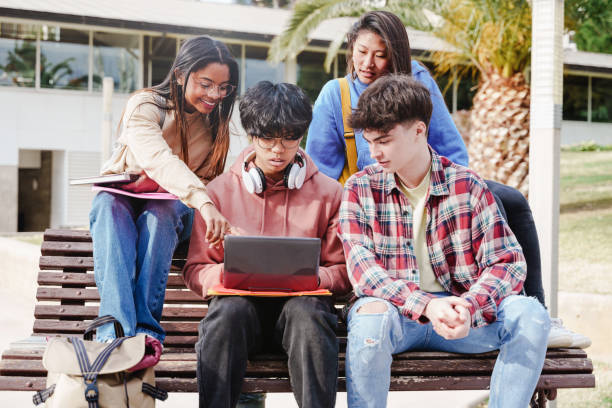 Four diverse students sitting on a bench, gathered around a laptop, collaborating and studying outdoors.