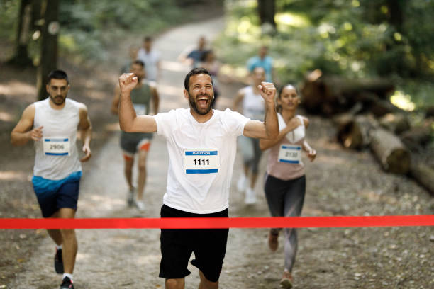 Professional Runner Competing In National Championship. A strong Caucasian Male Athlete is Sprinting Towards the Line in a Crowded Stadium, Crossing the Race Finish with His Hands in the Air