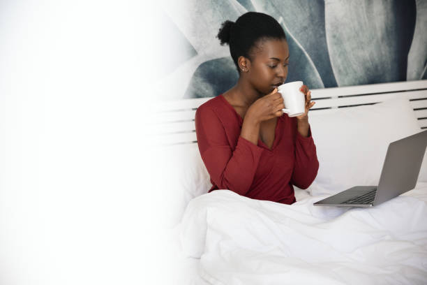 African woman sitting in bed sipping on coffee looking at laptop
