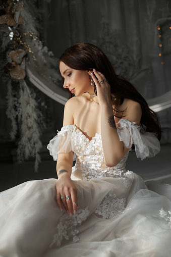 Woman Bride in a white dress is sitting on the floor by the window. A romantic image of a woman with a tattoo on her arm. Wedding