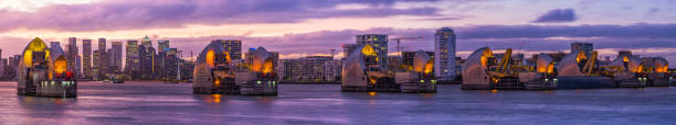 The gates of the Thames Barrier illuminated at sunset overlooked by the skyscrapers of Canary Wharf and Docklands along the River Thames in London, UK.