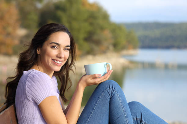 Happy woman holding coffee mug looks at camera in nature stock photo