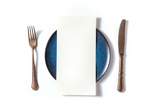 Dinner menu or invitation concept. A piece of paper on a table, with a fork, a knife, and a blue plate, overhead flat lay shot on a white background