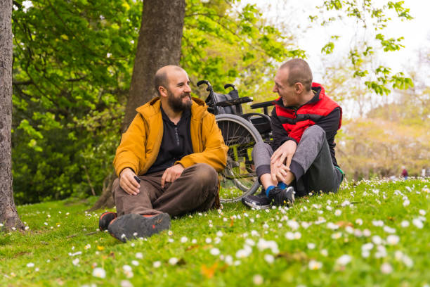 un joven paralizado con un amigo sentado en el césped de un parque público de la ciudad, hablando y riendo - physical impairment wheelchair disabled accessibility fotografías e imágenes de stock