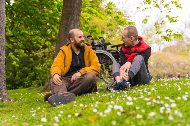 A paralyzed young man with a friend sitting on the grass of a public park in the city, talking and laughing stock photo