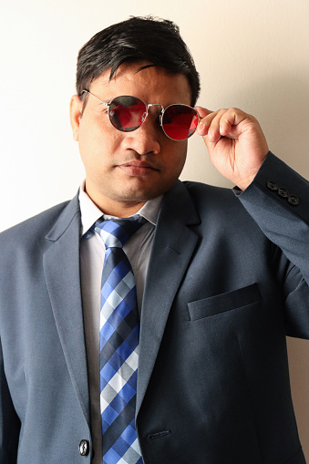 Stock photo showing three quarter length portrait of Indian business man wearing smart, blue suit, collared shirt and tie whilst removing mirrored sunglasses.