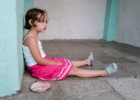 Cute angry girl is sitting on the floor near big stone. Concept of children behavior problem.