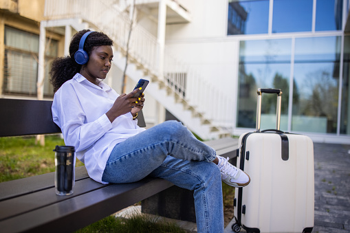 Young woman Black ethnicity listening to music, using mobile phone, while sitting on the bench, waiting for her transportation