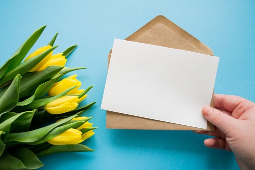 Hand holds envelope and paper for text on blue background and yellow tulips. Happy holiday greetings.
