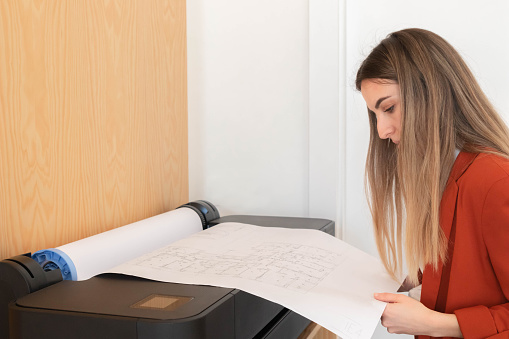 Young blonde woman in architecture, intrior design and engineering office printing blueprints in plotter