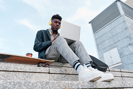 View from below of a young African-American adult man on a skateboard while working on a laptop computer