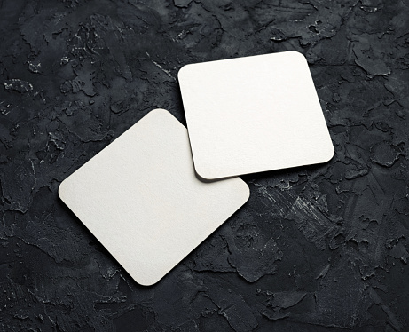 Two blank square beer coasters on black plaster background.