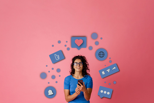 Bigger picture of a young female adult looking at the camera while stood in-front of a pink background. She is holding a mobile smart phone. A series of infographics are floating around her head to interpreted what she's doing on the phone.