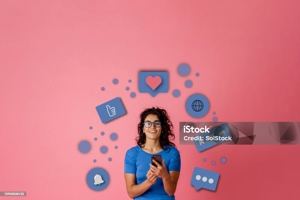 Connecting With Friends Online Bigger picture of a young female adult looking at the camera while stood in-front of a pink background. She is holding a mobile smart phone. A series of infographics are floating around her head to interpreted what she's doing on the phone. Advertisement Stock Photo