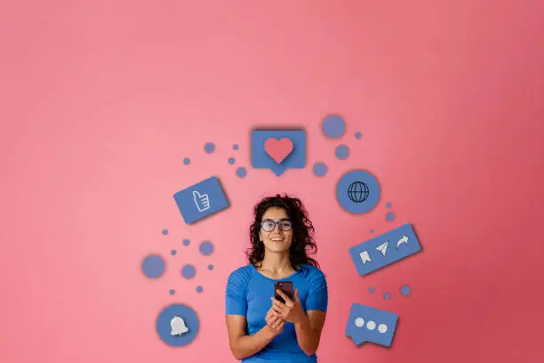 Bigger picture of a young female adult looking at the camera while stood in-front of a pink background. She is holding a mobile smart phone. A series of infographics are floating around her head to interpreted what she's doing on the phone.