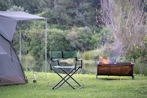 Campsite along river with campfire, canvas tent and camping chair. Outdoor leisure campground in nature.
