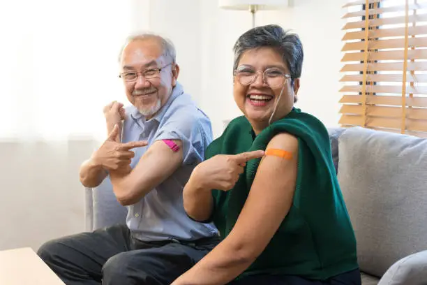 Photo of Asian couple get vaccinated with bandage on arm show thumb up sign in living room.